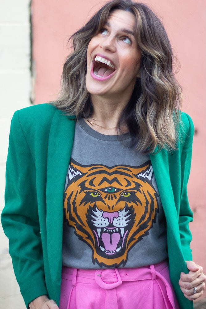 Woman wearing a green blazer over a gray t-shirt with a tiger graphic, standing against a pink wall.