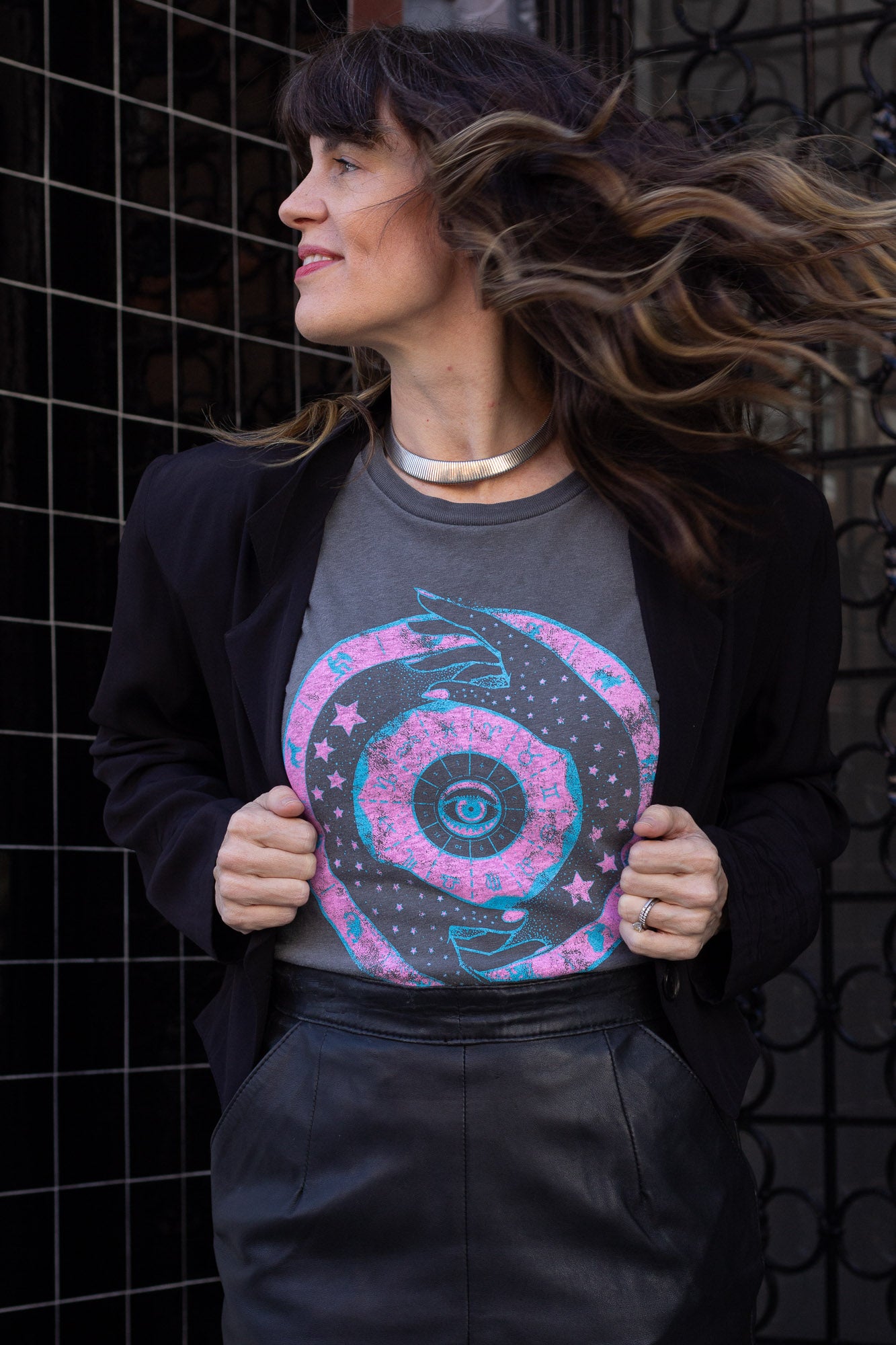 Woman wearing a gray t-shirt with a colorful zodiac design, standing against a black tiled wall.
