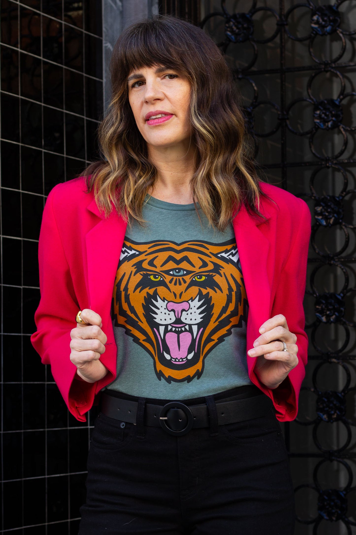 Woman wearing a pink blazer over a green graphic t-shirt with a tiger design, standing against a dark textured wall.