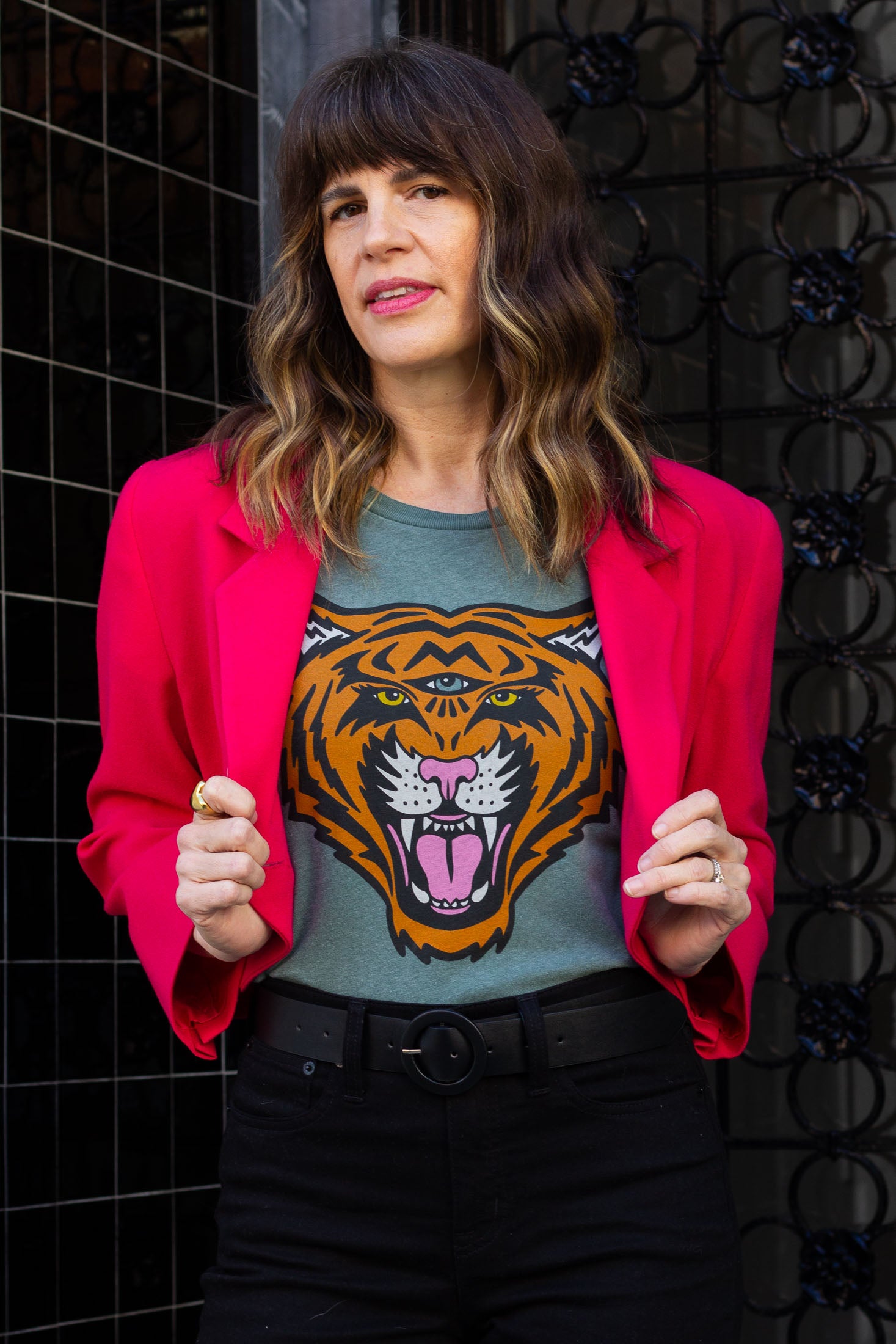 Woman wearing a pink blazer over a green graphic t-shirt with a tiger design, standing against a dark textured wall.