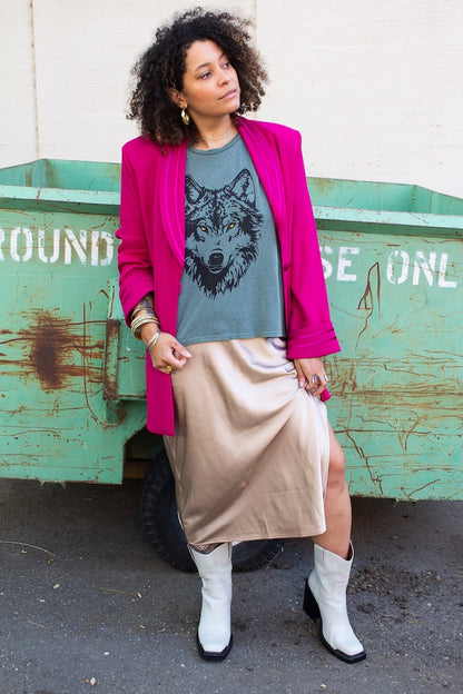Woman wearing a pink blazer, graphic t-shirt, beige skirt, and white boots standing in front of a green dumpster.