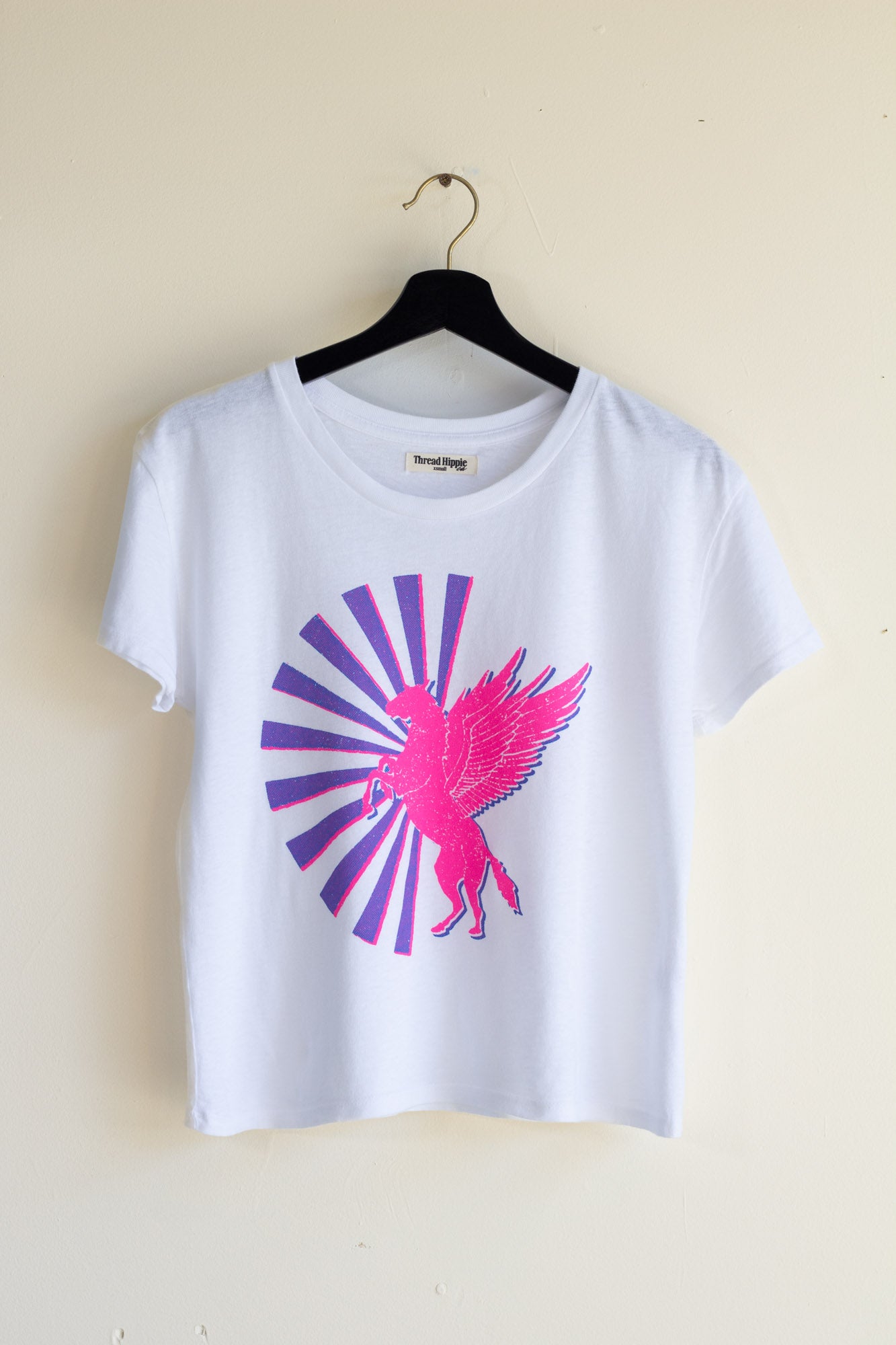 Woman's white t-shirt with pink and purple graphic design on a hanger against a beige wall.