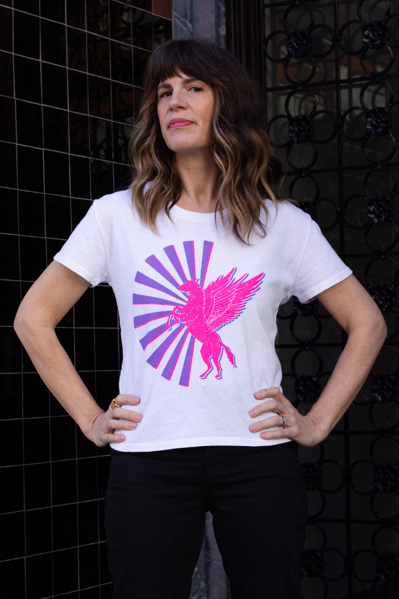 Woman wearing a white t-shirt with a pink and purple horse graphic design against a black tiled wall.