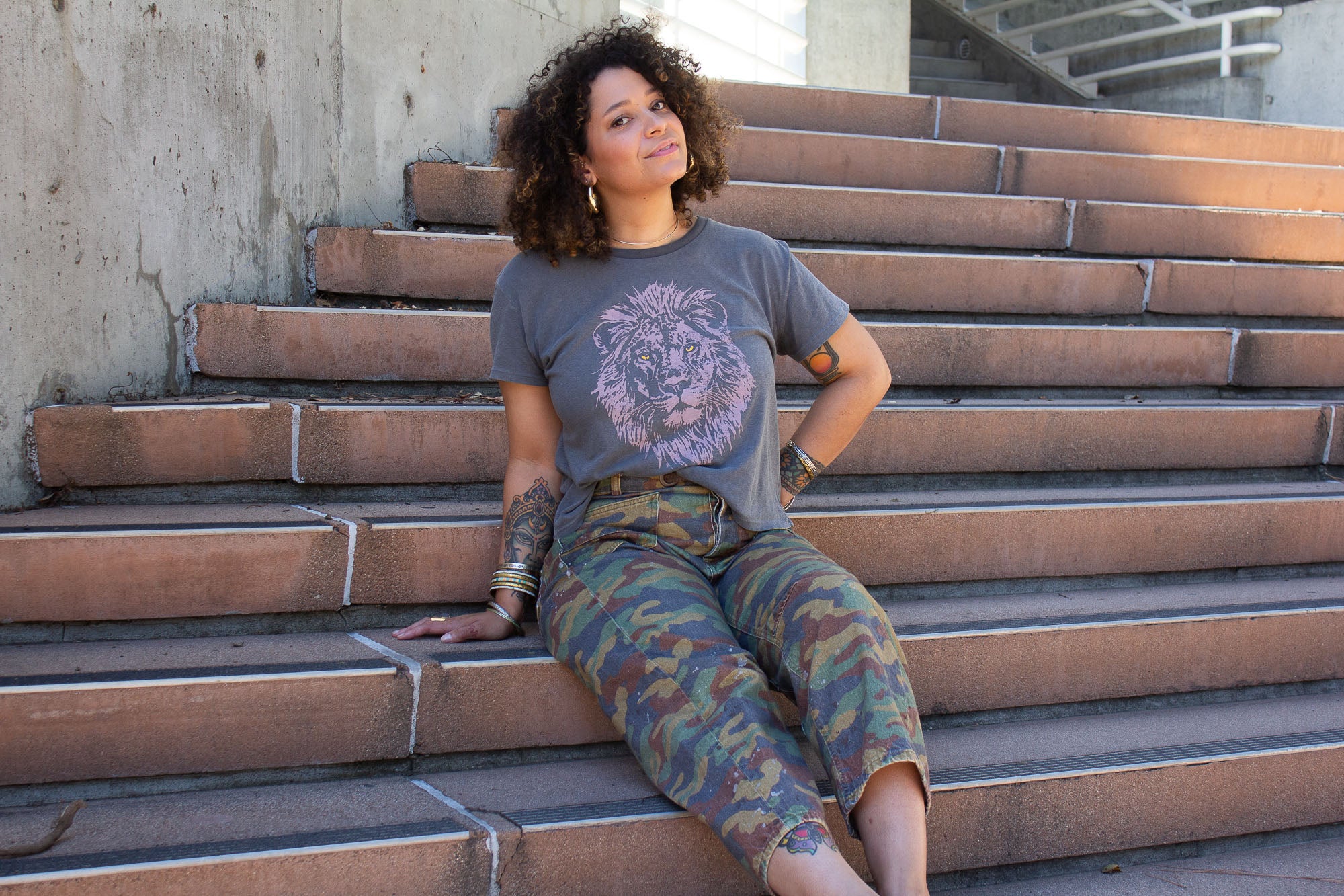Woman sitting on outdoor steps wearing a lion graphic t-shirt and camouflage pants.