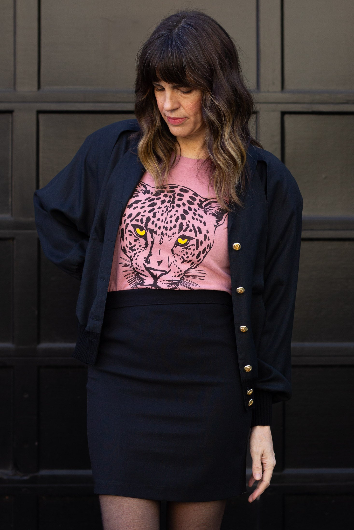 Woman wearing a black blazer over a pink t-shirt with a leopard graphic, standing against a dark background.