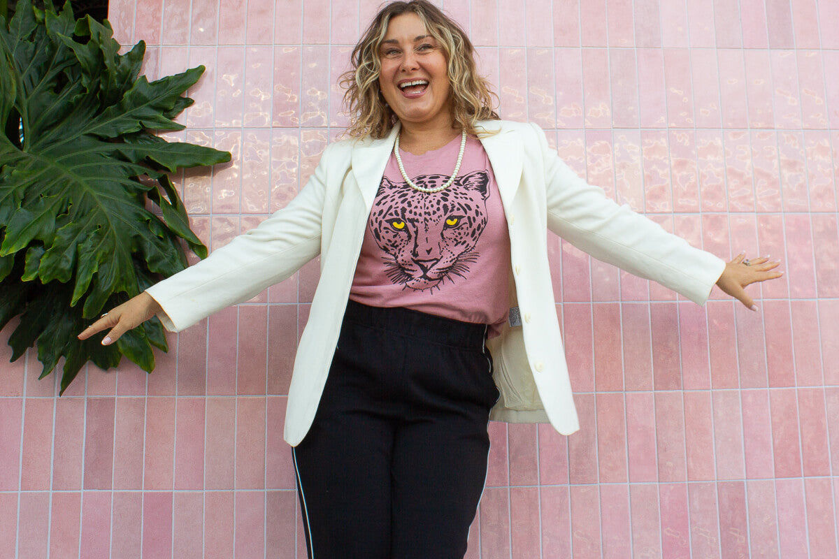 Woman wearing a white blazer, pink shirt with a leopard print, and black pants against a pink tiled wall.