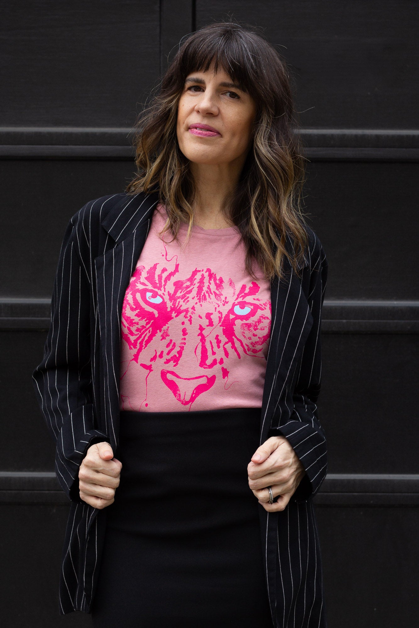 Woman wearing a pink t-shirt with a tiger design and a black pinstripe jacket against a black background.
