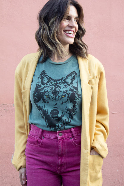 Smiling woman wearing a green tee with a wolf graphic, yellow cardigan, and pink pants against a pink wall.