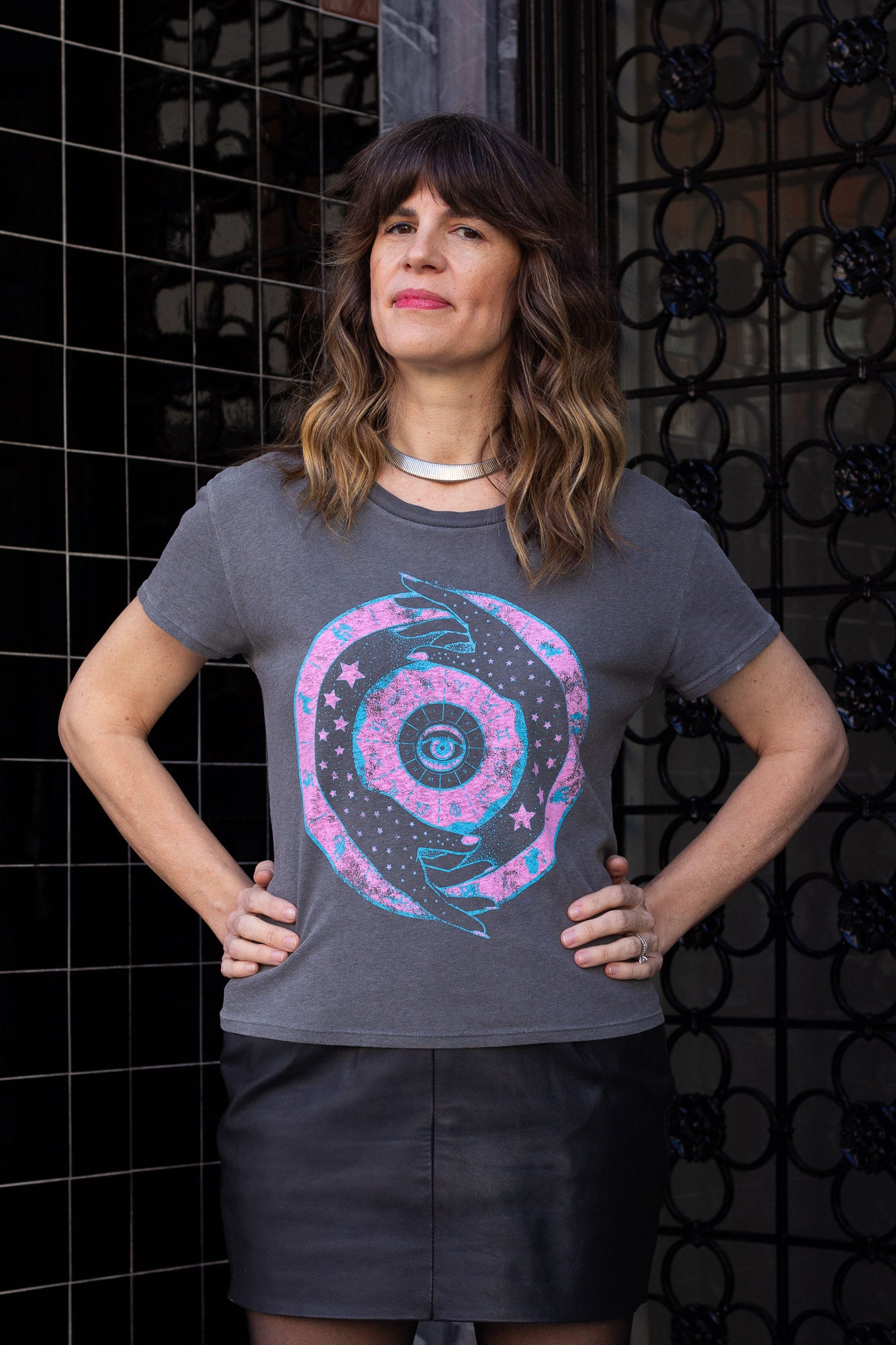 Woman wearing a gray t-shirt with a zodiac design against a black tiled wall.