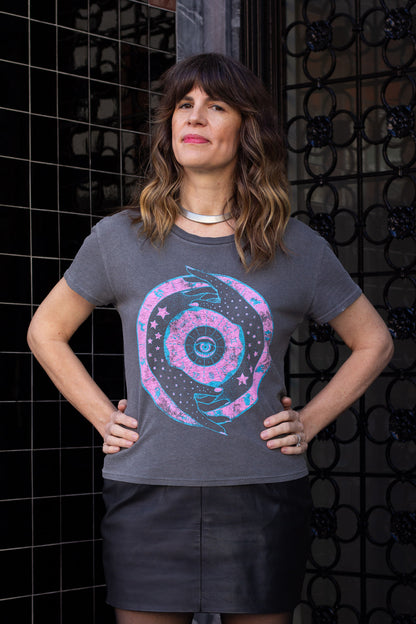 Woman wearing a gray t-shirt with a zodiac design against a black tiled wall.
